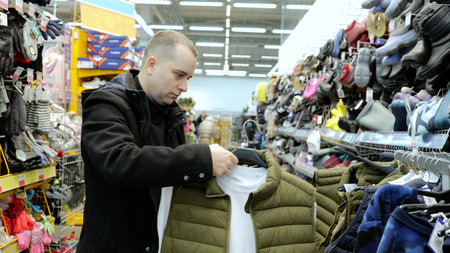 Male shopper comparing white t shirt and green vest on hangers, browsing clothing selection within crowded retail environment of modern shopping centerの写真素材