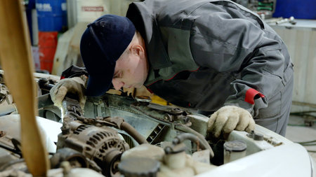 Experienced auto mechanic wearing gloves and uniform carefully inspecting car engine compartment in a professional garage workshop, performing diagnostics and maintenanceの写真素材