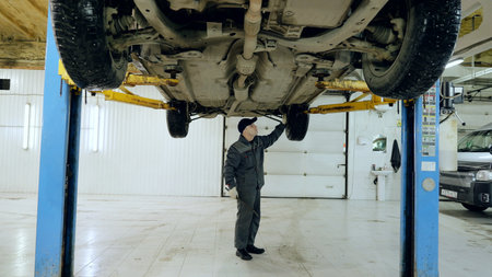 Skilled mechanic inspecting vehicle underside on hydraulic lift, checking mechanical components in modern automotive workshop repair environmentの写真素材