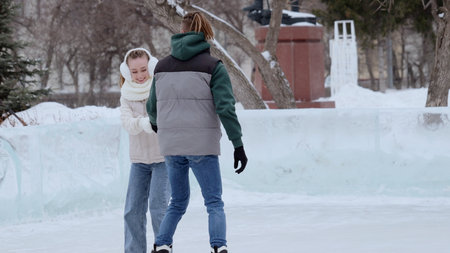 Young couple holding hands, gliding across ice rink in snowy winter park, sharing intimate moment...