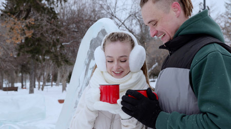 Loving couple drinking hot beverages, bundled in winter gear near frosty ice sculpture, sharing warm...