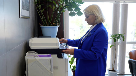 Efficient businesswoman in a blue suit using a multifunction printer in a modern office surrounded...