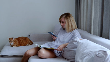 Middle aged woman enjoying leisure time at home, sitting on a comfortable sofa with her ginger cat,...
