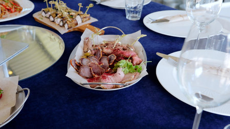 Charcuterie plate with sliced cured meats, garnished by fresh parsley, resting on metal tray with parchment, near empty wine glasses on blue linenの写真素材