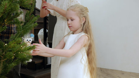 Young girl with golden hair placing owl ornament on christmas tree, mother hanging decorations behind her in festive living room settingの写真素材