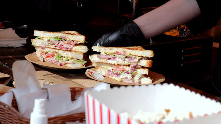 Vendor crafts multitiered deli sandwiches, Food stand displays stacked sandwiches with condiments and popcorn, Bustling street market scene with vendor preparing multilayer sandwiches and snack boxesの写真素材