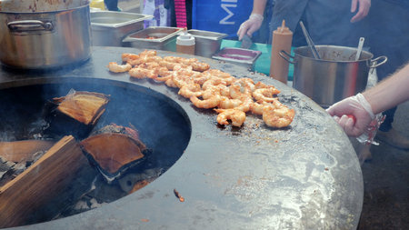 Shrimp sizzling on flat griddle with vendor flipping prawns using spatula, bubbling oil and aromatic spices, outdoor market stall with pots and condiments in background and casual crowd nearbyの写真素材