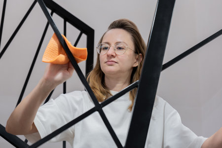 young woman cleaning glass mirror with orange cloth, wearing white shirt and glasses, focused...