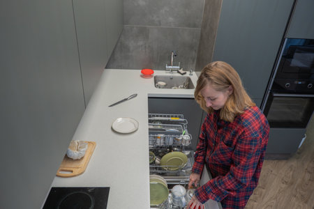 Young white woman arranging plates inside dishwasher for styling shot, captured from overhead in contemporary kitchen, focused expression, red plaid shirt, neat racks, ceramic plates and glasswareの写真素材
