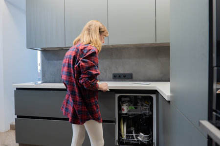 Young white woman loading dishwasher in contemporary gray kitchen, wearing red plaid shirt, focused on placing glasses and plates into lower rack, minimalist cabinets and stainless countertop, naturalの写真素材