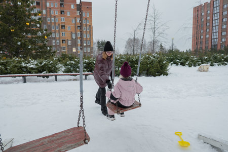 Woman pushing toddler on snowy swing captured mid-push, child in pink snowsuit with rubber boots, mittens and knit hat, adult wearing puffer jacket and beanie, shovel toy visible on snow, urbanの写真素材