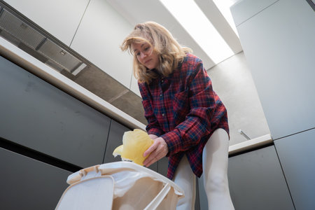 A woman throws trash into a trash can in a neat modern kitchen while cleaning up after cooking. The pose is casual, demonstrating everyday household chores.の写真素材