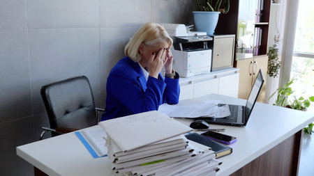 Senior businesswoman suffering from headache and eye strain after working too long on computer in office, massaging her temples and eyes, sitting at desk with laptop and paperworkの写真素材