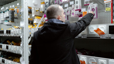 Skilled electrician reviewing light switches and electrical fixtures in hardware store, comparing...