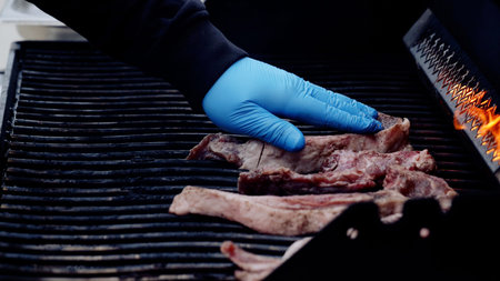 Professional chef wearing blue gloves grilling succulent ribs, flames licking meat on barbecue grill, preparing savory meal with culinary expertise