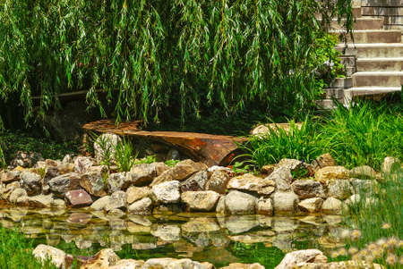 Wooden garden bench in the shape of half a circle by a rocky pond surrounded by greeneryの写真素材