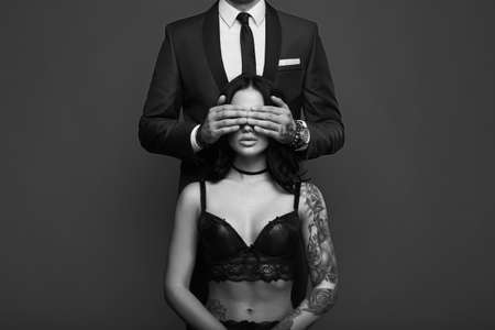 Portrait of a brutal man in elegant suit touches sexy girl with a tattoo in lingerie on gray backgroundの写真素材