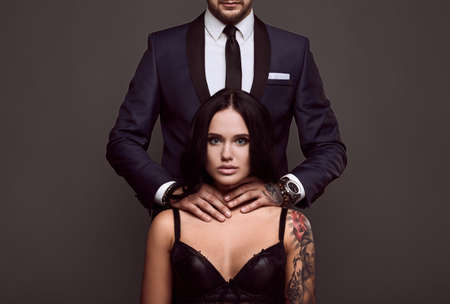 Portrait of a brutal man in elegant suit touches sexy girl with a tattoo in lingerie on gray backgroundの写真素材