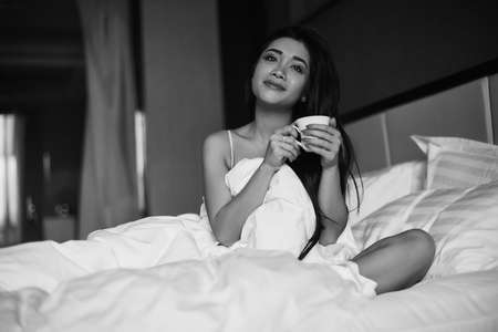 Portrait of a beautiful sensual asian woman sitting on a bed under a blanket with a cup of coffee in hands. Interior shotの写真素材