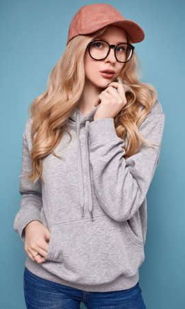 Portrait of a cute positive blonde woman in a pink baseball cap, glasses and sweatshirt on bright...