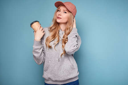 Portrait of a cute positive blonde woman in a pink baseball cap, cup of coffee and sweatshirt on bright background in studioの写真素材