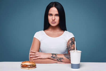 Glamorous fashion portrait of beautiful charming tattoo girl with hamburger and soda on a bright...