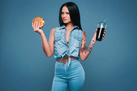 Glamorous fashion portrait of beautiful charming tattoo girl with hamburger and soda on a bright background in the studioの写真素材