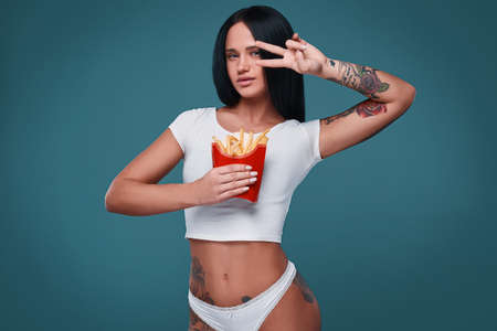 Glamorous fashion portrait of beautiful charming girl with tattoo holding a french fries on a bright background in the studioの写真素材