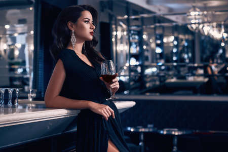 Gorgeous beauty young brunette woman in dark dress with glass of wine standing at the bar in luxury interiorの写真素材