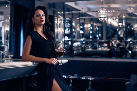 Gorgeous beauty young brunette woman in dark dress with glass of wine standing at the bar in luxury interiorの写真素材