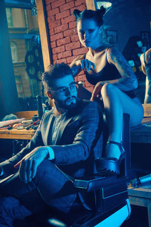 Colorful portrait of beautiful couple: brutal man in elegant suit and sexy girl with a tattoo wearing lingerie in barbershopの写真素材