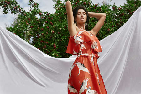 Portrait of gorgeous brunette woman in fashion orange dress posing among an apple trees
