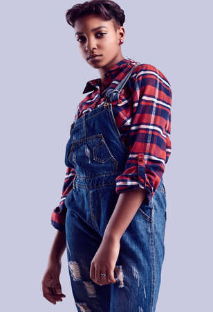 Sensual portrait of glamor elegant black hipster girl with short hair in a plaid shirt and denim overalls posing on colorful background in studioの写真素材