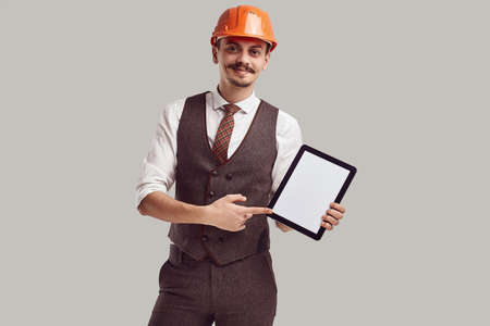 Portrait of handsome young confident arabic architect with fancy mustache in woolen suit and construction helmet holds tabletの写真素材