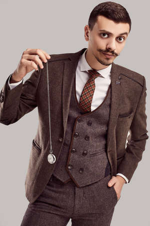 Portrait of handsome young confident arabic businessman with fancy mustache in woolen brown full suit holds antique clock on studio backgroundの写真素材