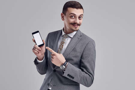 Portrait of handsome young confident arabic businessman with fancy mustache in fashion gray full suit points to the phone on studio backgroundの写真素材