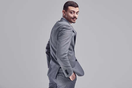 Portrait of handsome young confident arabic businessman with fancy mustache in fashion gray full suit looks over his shoulder on studio backgroundの写真素材