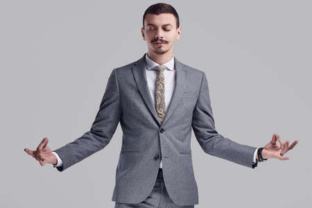 Portrait of handsome young confident arabic businessman with fancy mustache in fashion gray full suit meditating on studio backgroundの写真素材