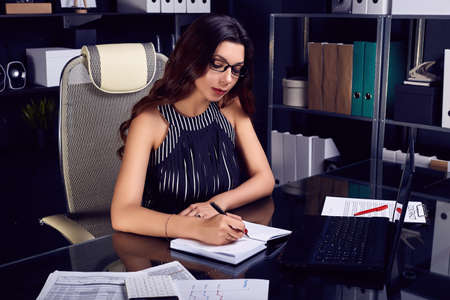 Portrait of young beautiful business woman in fashion suit and glasses working at stylish black computer deskの写真素材