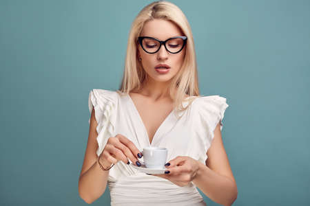 Portrait of gorgeous sensual blonde woman in fashion white dress and glasses with cup of coffee posing on blue background in studioの写真素材