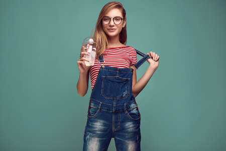 Portrait of cute teenage girl in denim overalls and glasses with soda water posing on color background. Studio shoot.