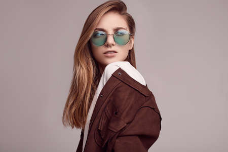 Portrait of cute teenage girl in fashion jacket and color glasses posing on gray background. Studio shoot.の写真素材