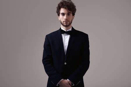 Fashion portrait of handsome elegant man with curly hair wearing tuxedo posing on gray background in studioの写真素材