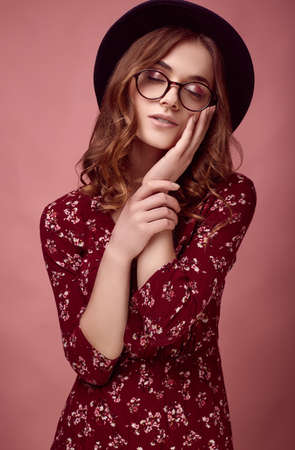 Sensual portrait of elegant glamor hipster girl in red fashion dress, black hat and glasses posing on colorful pink background in studioの写真素材