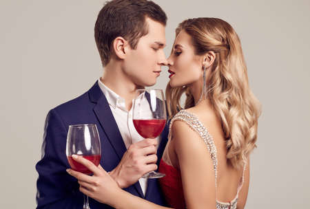 Portrait of sensual beautiful young couple dressed in formal clothes: woman in fashion luxury red dress and man wearing blue classical suit with glasses of wine isolated on white background.の写真素材