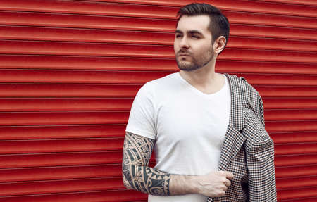 Portrait of young handsome brutal man with tattoo in a fashion suit stands near the red metal gate. Street style.の写真素材
