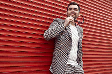 Portrait of young handsome brutal man in a fashion suit and sunglasses stands near the red metal gate. Street style.の写真素材