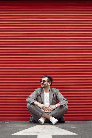 Portrait of young handsome brutal man in a fashion suit and sunglasses with cup of coffee sitting next to the red metal gate.の写真素材