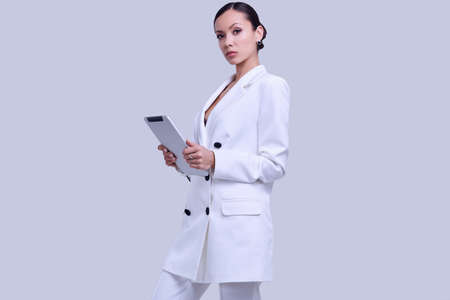 Portrait of a cute and gorgeous latin women in fashion white suit with digital tablet posing on bright studio backgroundの写真素材