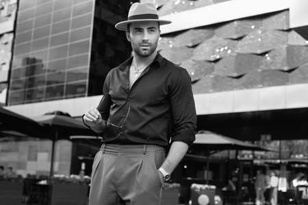 Portrait of a beautiful brutal tanned muscular hipster man in black shirt, hat and sunglasses posing in the streets of the European cityの写真素材